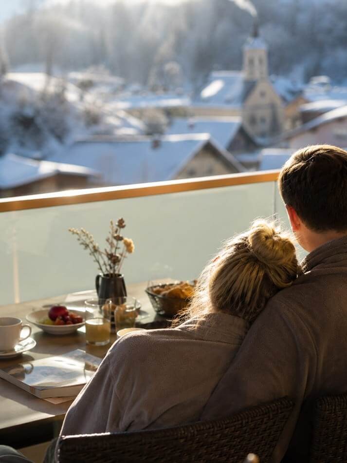 Paar genie&szlig;t entspannte Wohlf&uuml;hlmomente beim Fr&uuml;hst&uuml;ck auf dem Balkon mit Blick auf das winterliche Wagrain