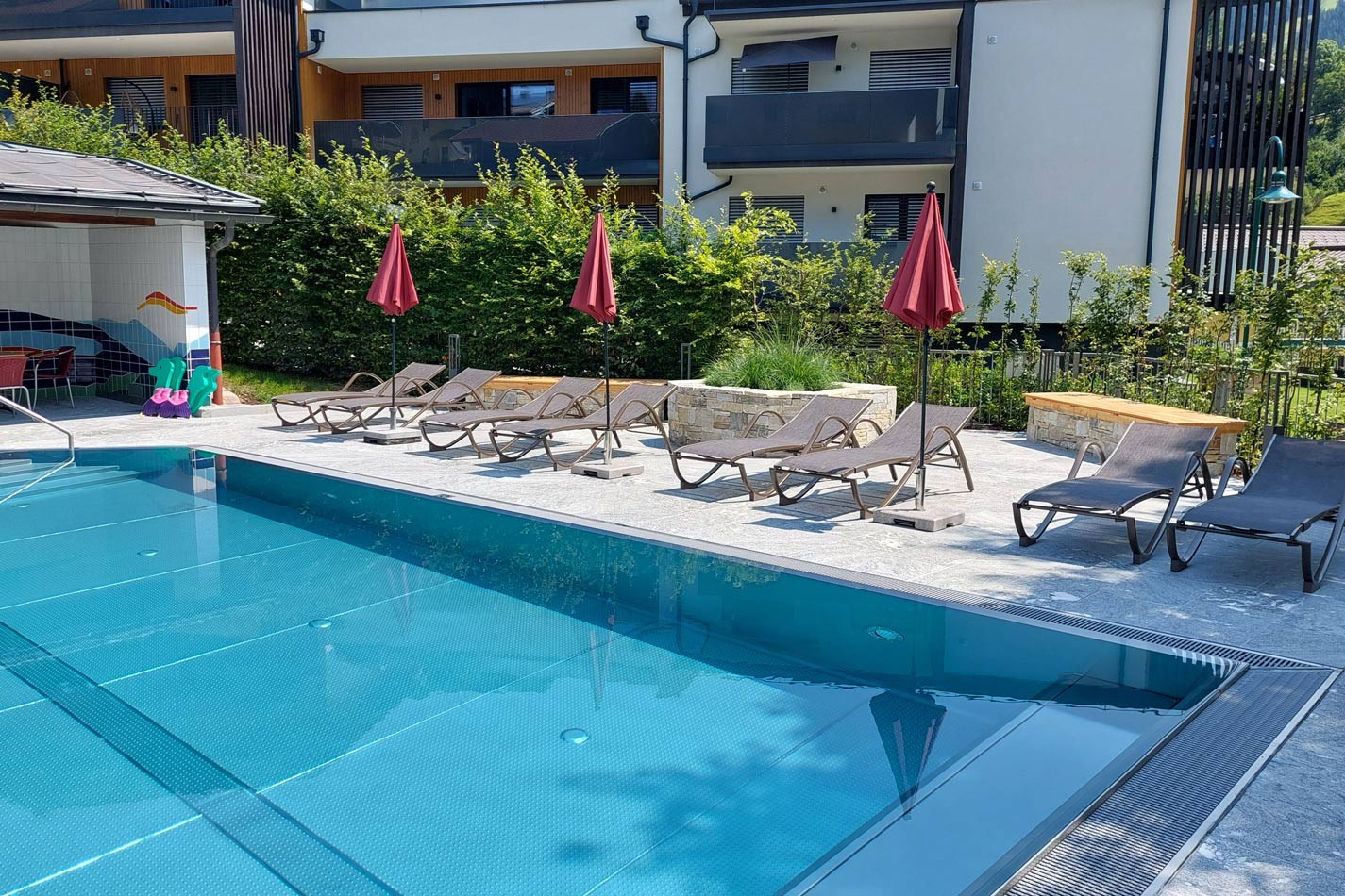 Poolbereich mit Liegest&uuml;hlen und roten Sonnenschirmen vor einem Geb&auml;ude